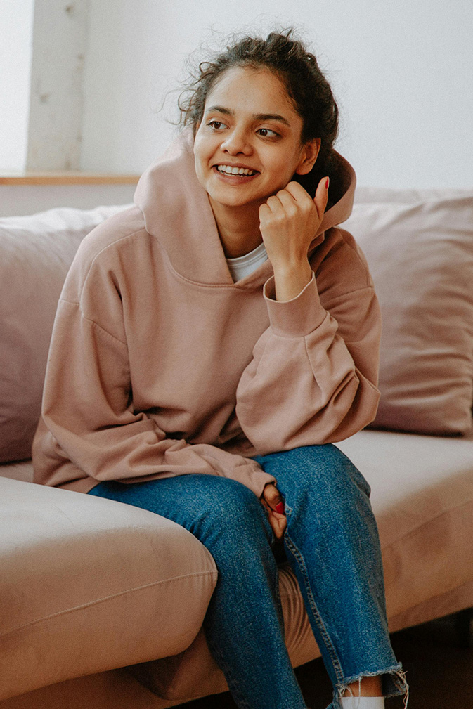 A young woman in a hoodie and jeans sits smiling on a beige couch